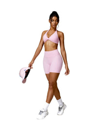 Woman wearing pink ORU Mika Short and matching sports bra holding a pink cap, posing in athletic shoes and white socks
